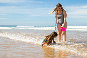 Fototapeta premium Joyful mom and little daughter standing ankle deep in sea water and wet sand, picking shells into bucket. Bright blue sea with waves and white foam in background. Family summer holidays concept