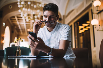 Smiling young man taking off eyeglasses while using smartphone in cafe
