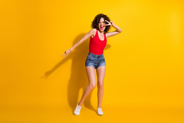 Full length body size photo of pretty female hipster with curly hairdo dancing moving smiling showing gesture v-sign with two fingers at party isolated on vivid yellow color background