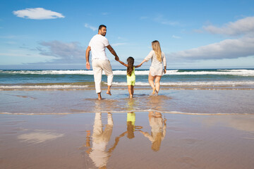 Family couple and kid walking ankle deep in sea water, spending leisure time on beach. Rear view. Family outdoor activities concept