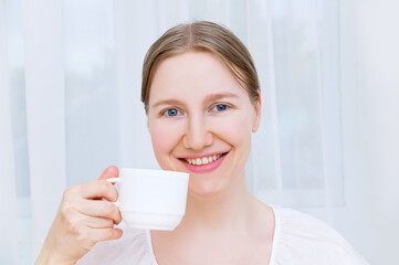 Caucasian blond woman sitting at a table smiling cheerfully holding  white cup of coffee in her hands.
