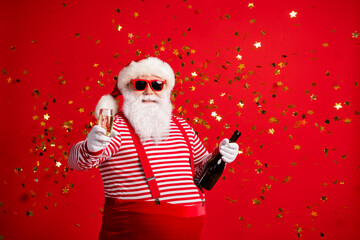 Photo of grandpa grey beard hold bottle wine cheerful drink alcohol smile wear santa claus x-mas costume suspenders sunglass striped shirt cap isolated red color background