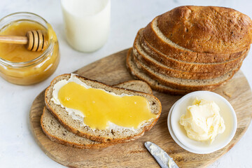 Slice of sourdough bread with honey, butter and milk on wooden chopping board, white table. Healthy, organic country breakfast with natural morning light. Copy space. Freshly baked bread.