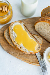 Slice of sourdough bread with honey, butter and milk on wooden chopping board, white table. Healthy, organic country breakfast with natural morning light. Copy space. Freshly baked bread.
