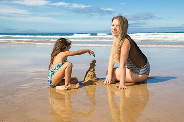 Cheerful mom and little daughter building sandcastle on beach, sitting on wet sand, enjoying vacation at sea. Family summer holidays concept