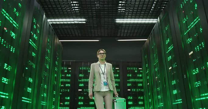 Caucasian woman technician with tools box coming in and walking among servers of big data information. Green working processors in database storage. Female engineer worker of telecommunication.