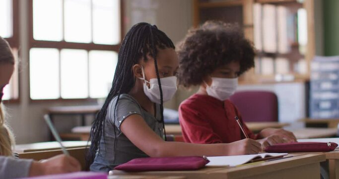 African American girl group of kids wearing face masks writing while sitting on their desk at school