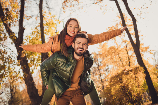 Low angle view photo of bonding couple man carry piggyback girl hold hands in autumn september city park wear coats
