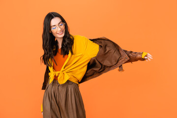 stylish woman in glasses and leather jacket standing with outstretched hand isolated on orange