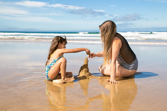 Little Girl And Her Mom Building Sandcastle On Beach, Sitting On Wet Sand, Enjoying Vacation At Sea. Family Summer Holidays Concept