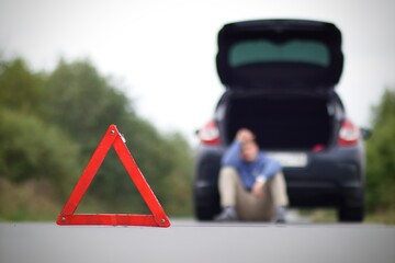 Accident on the road. Emergency warning triangle and a blurred man sitting near damaged car with open trunk