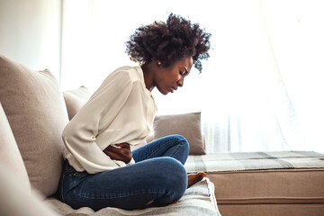 View of young woman suffering from stomachache on sofa at home. Woman sitting on bed and having...