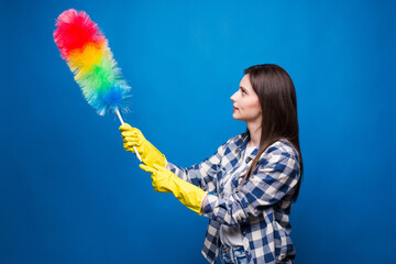 Portrait of optimistic housewife wearing yellow rubber gloves holding colorful duster while doing housework and cleaning room isolated over blue background