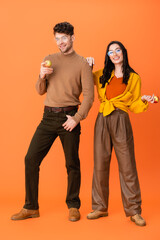 Obraz premium full length of trendy couple in glasses and autumn outfit holding apples on orange