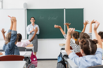 Fototapeta back view of multicultural pupils with hands in air, and teacher standing at chalkboard with back to school lettering obraz