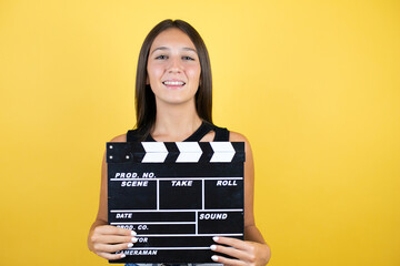 Beautiful young girl kid over isolated yellow background holding clapperboard very happy having fun