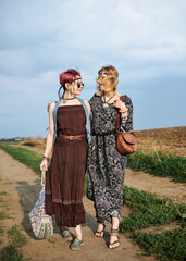 Two hippie women, wearing boho style clothes, walking on dirt road on green field, having fun. Female friends, traveling together in countryside. Eco tourism concept. Summer leisure free time.