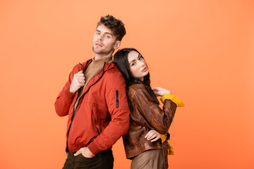 trendy couple in leather jackets looking at camera while posing isolated on orange