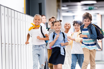 selective focus of excited multicultural classmates with backpacks running along school corridor