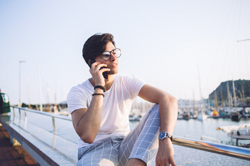 Handsome 20s male traveler in trendy wear resting outdoors near bay in city making smartphone call, young hipster guy in stylish outfit and spectacles enjoying free time and mobile phone talk