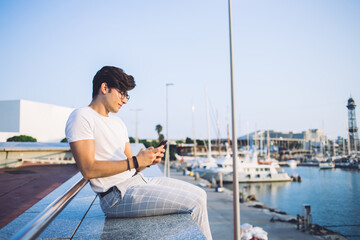 Fototapeta na wymiar Young trendy dressed caucasian male traveler recreating on free time near harbor share photo in social networks, pensive caucasian hipster guy reading message and sending text recreating outdoors