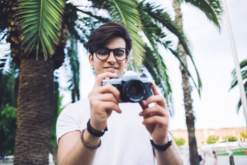 Cheerful male in eyewear holding camera making photo during trip in town explore city on sunny day, smiling caucasian man traveler satisfied with journey taking photos of town standing near palm trees