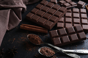Tasty milk chocolate and spoons with cocoa powder on black table