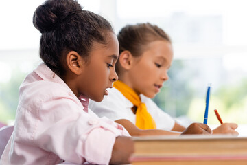 Selective focus of african american schoolgirl holding pen near friend and books in classroom