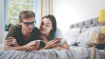 Lovely young happy couple laying on bed using mobile phones and talking