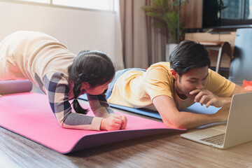 The Asian young man lay down on yoga mat with laptop and children near by while do planking in the living room together. Family spend time together at home.