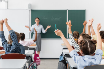 back view of multicultural pupils with hands in air, and teacher standing at chalkboard with open...