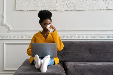 Afro-American biracial woman with afro hairstyle in yellow cardigan sitting on couch, working online on laptop, watching webinar or videos in social media, holding mug, enjoying drinking coffee