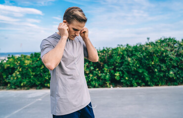 Pensive caucasian male jogger putting on earphones for listening music during cardio workout, serious sportsman enjoying audiobook via accessory for motivating on training outdoors keeping fit