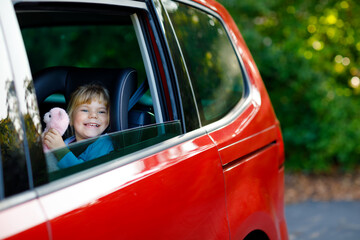 Adorable toddler girl sitting in car seat, holding plush soft toy and looking out of the window on...