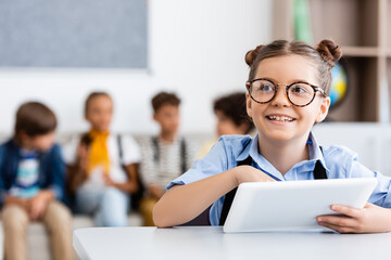 Selective focus of schoolgirl with digital tablet sitting at desk near friends at background in classroom