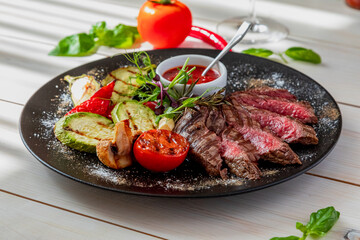 chopped medium rare steak with grilled vegetables. Sauce is served with the dish. Dish on a white plate in natural light.