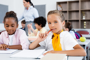 Selective focus of schoolgirl looking at camera near african american friend at desk in school