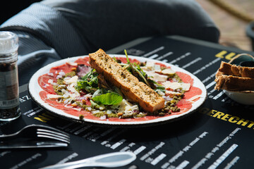 A plate with Beef Carpaccio, with cheese, lettuce, pine nuts, delicious with a sandwich, stands on a terrace table, people eat and drink in the background