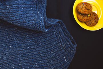 Blue knit sweater and yellow plate with flavored cookies with colored drops on black background. Craft on black background. Texture beautiful knitted fabric as background. Female hobby concept