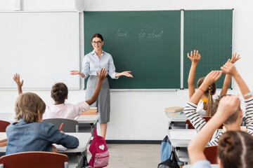 back view of multiethnic pupils with hands in air, and teacher standing with open arms near...