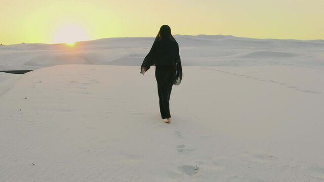 A silhouette woman in a black oriental dress walking in the desert, rear view. The head is covered with a scarf, Long dress flying in wind. Arabic nature background, white sands, dramatic sky, sunset