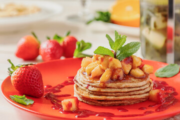 pancakes with jam and strawberry pieces. The dessert is decorated with a sprig of mint. There are tea, juice, an orange slice and a strawberry on a white background. In natural light