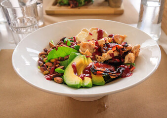 Delicious and healthy lunch salad: round plate lettuce, avocado, bacon, chicken, pistachio nuts, and a pink sauce, served with water on a brown paper place mat.