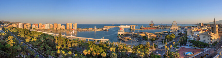 Malaga, Spain: seafront and harbour at sunset  with ferris wheel at the harbour entrance. In the background the harbour in full operation with a freighter and harbour cranes.