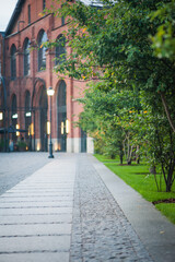 Obraz premium A cobblestone path leading to a large red brick building, a path that runs along green trees. Vertical photo.