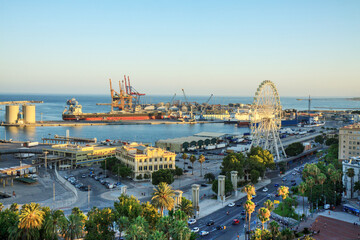 Malaga, Spain: seafront and harbour at sunset  with ferris wheel at the harbour entrance. In the background the harbour in full operation with a freighter and harbour cranes.