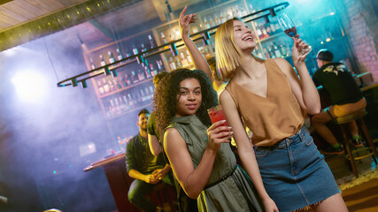 Attractive young women getting drunk, posing with cocktail in their hands. Friends celebrating, having fun in the bar
