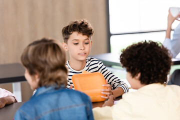 selective focus of schoolboy opening lunch box while sitting in school eatery near classmates