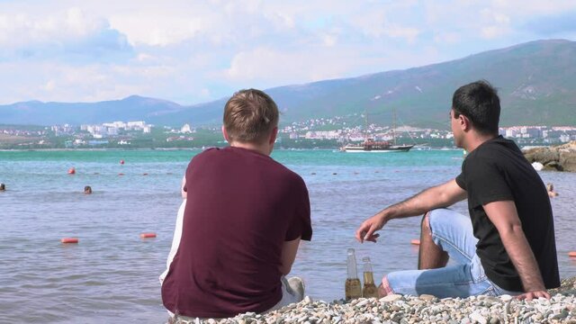 Two friends with beer sitting on beach by sea. Media. Two men sit with beer on beach on background of people swimming in sea. Vacation at sea with your best friend