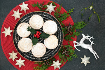 Delicious Christmas mince pies on a plate with winter berry holly, cedar cypress, mistletoe, silver star & reindeer decorations on grey grunge  background. Festive food composition. Top view.